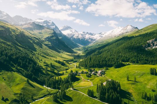 Luftaufnahme des Fextals im Sommer mit sattgrünen Wiesen, Bergwäldern, einem kleinen Weiler und schneebedeckten Gipfeln im Hintergrund