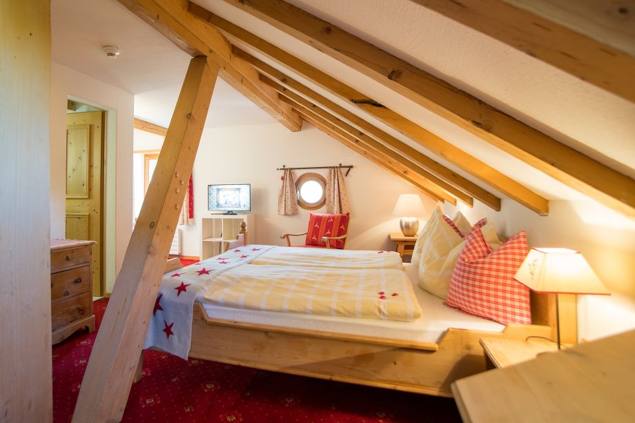 Gemütliches Dachzimmer der Sonne Fex mit sichtbaren Holzbalken, traditionellen Holzmöbeln, rotem Teppich und rot-weiss karierten Kissen