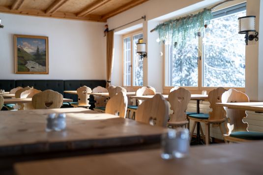Gemütlicher Speisesaal der Sonne Fex mit traditionellen Engadiner Holzstühlen, rustikalen Holztischen und Blick durch verschneite Bäume